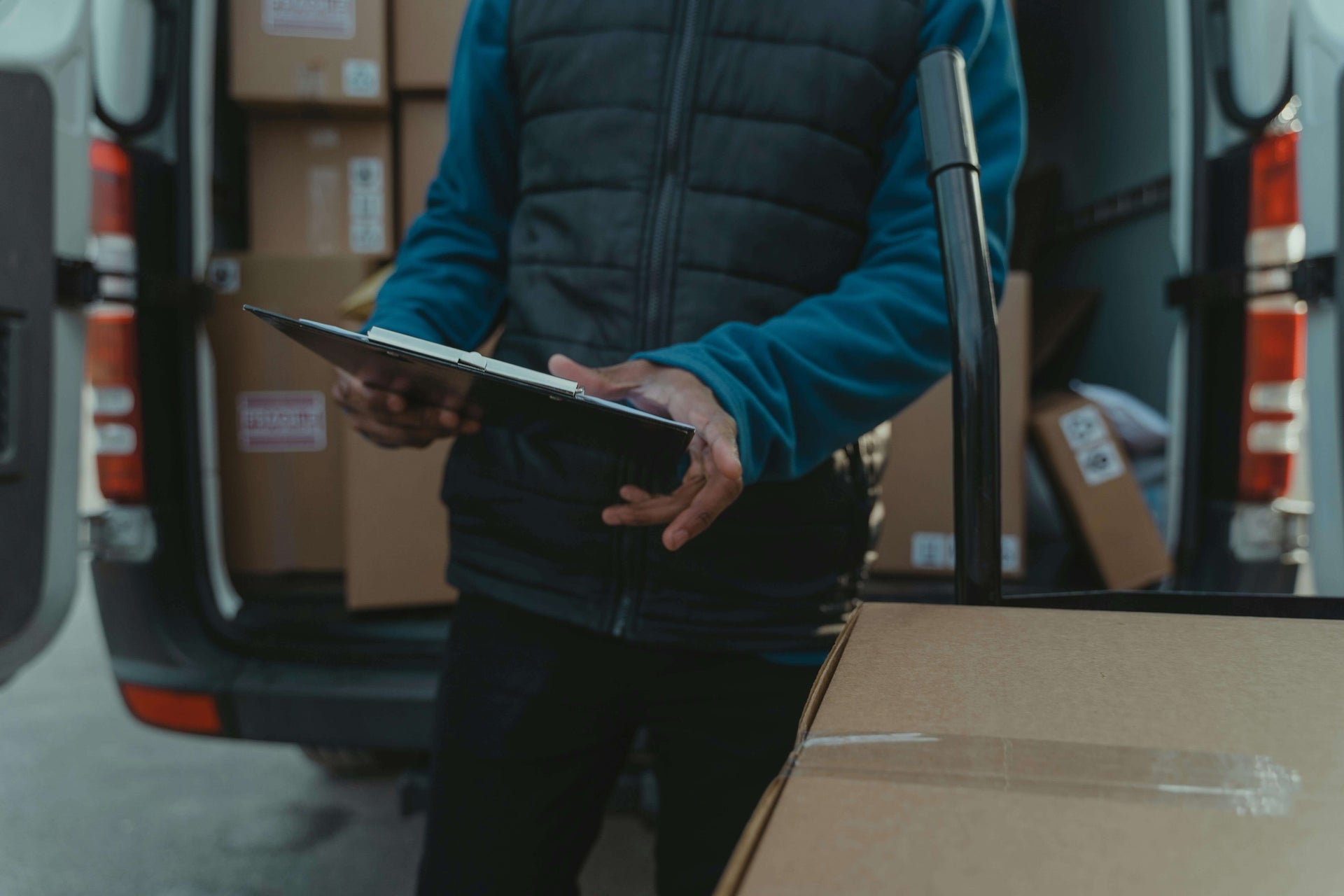 This image shows a delivery person standing next to an open van filled with packages. The individual is holding a clipboard, likely checking or signing off on deliveries, while another large box is on a dolly in the foreground. The scene captures the process of package delivery, emphasizing logistics, efficiency, and organization in a professional setting.