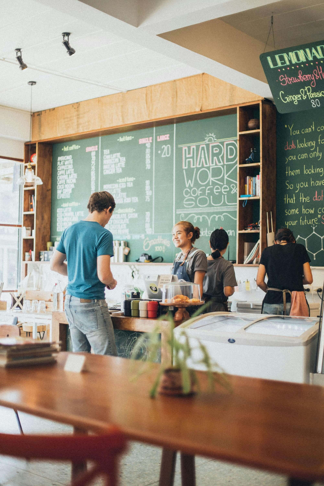 Stock image of inside a coffee shop. A customer is talking to the cashier, and the interior of the coffee shop is inviting and welcoming.