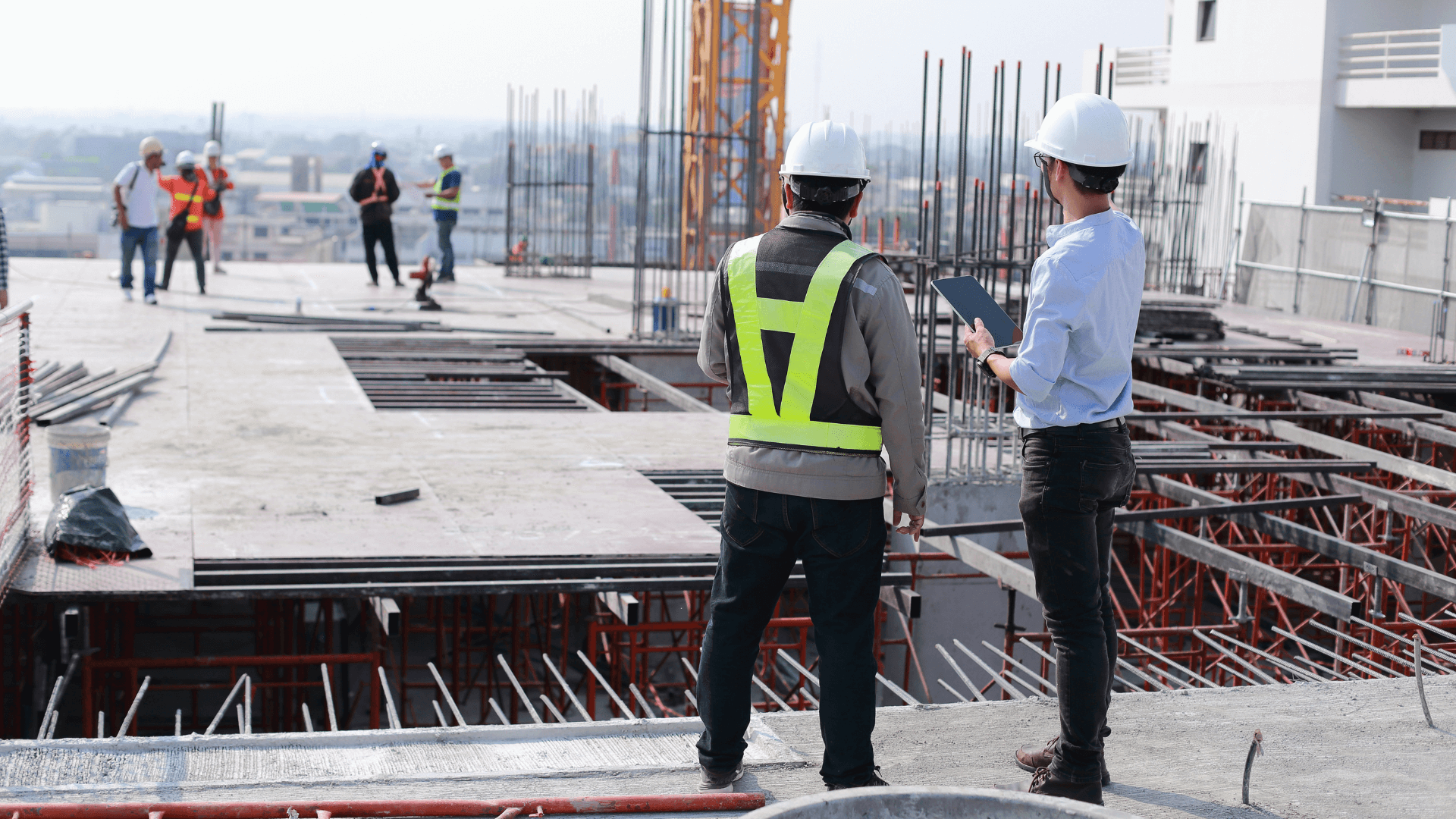 This image shows a construction site where multiple workers are engaged in various tasks. In the foreground, two men wearing hard hats and safety vests are standing and observing the site. One of them is holding a tablet, likely reviewing plans or data. The construction site appears to be on a building's upper level, with rebar and scaffolding visible, indicating that the structure is still in progress. In the background, other workers in hard hats and safety gear can be seen walking and working.