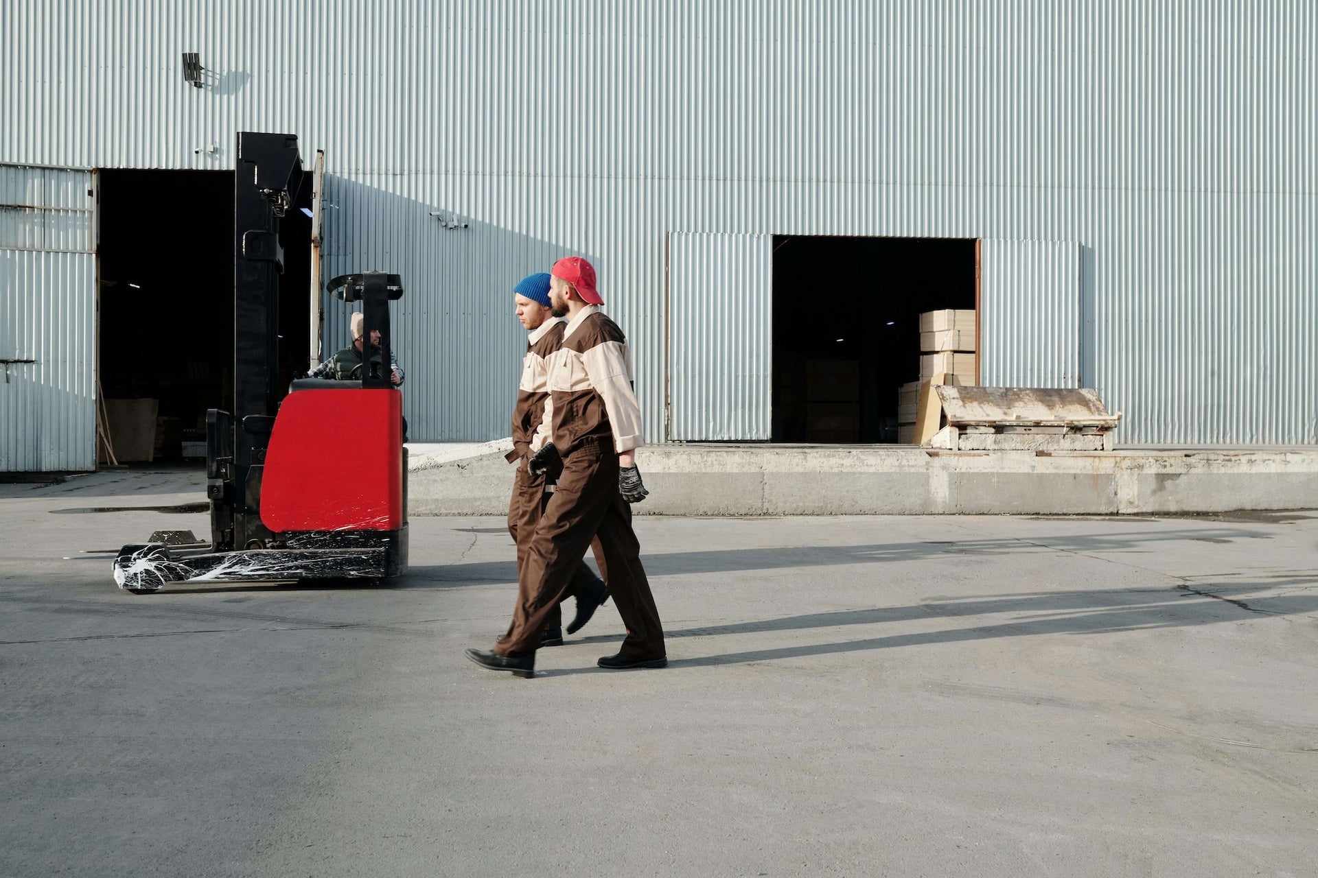 This image shows a warehouse or industrial setting where two workers, dressed in matching brown and beige uniforms and wearing beanies, are walking side by side outside the building. In the background, a forklift with a red body is being operated near a large open door, which reveals stacks of boxes inside the warehouse. The building has a corrugated metal exterior, with two large open doors leading to the interior. The overall scene suggests a busy distribution or logistics environment, with workers moving