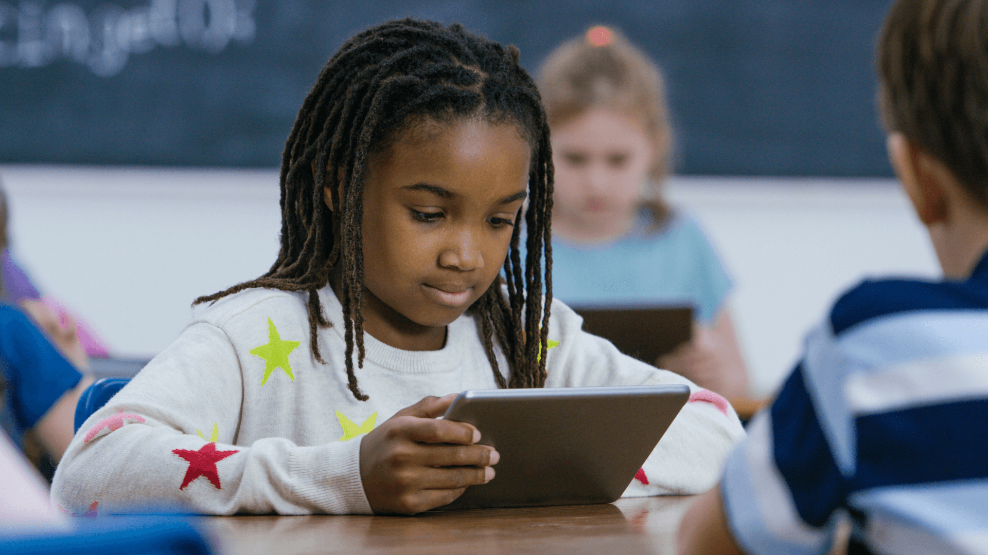 This image shows a young girl in a classroom setting, focused on using a tablet. She is wearing a sweater with colorful star patterns, and other children in the background are also engaged with their own tablets. The scene conveys a modern, tech-enhanced learning environment where students are using digital devices for education.