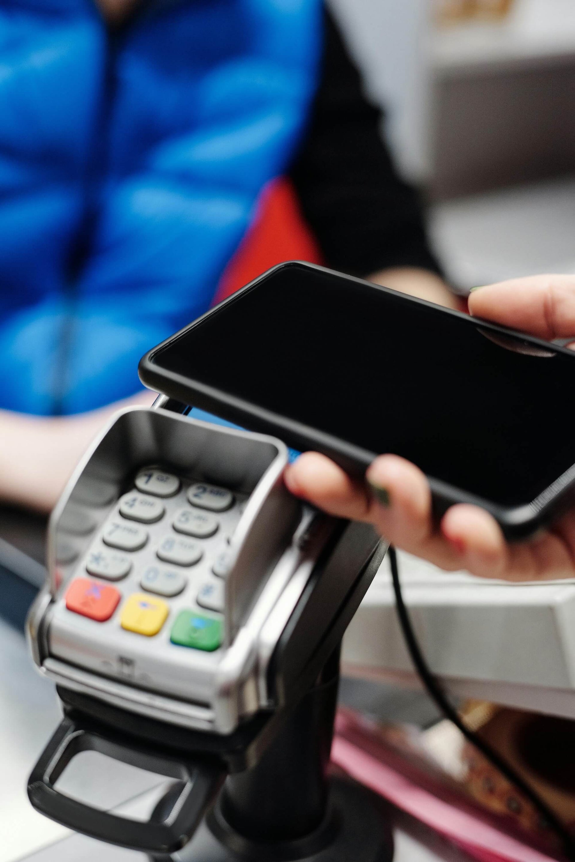 A person is using a smartphone to make a contactless payment on a card reader at a checkout. The close-up image focuses on the phone hovering over the payment terminal, with buttons and a screen on the device visible. The background is slightly blurred, emphasizing the transaction in progress.