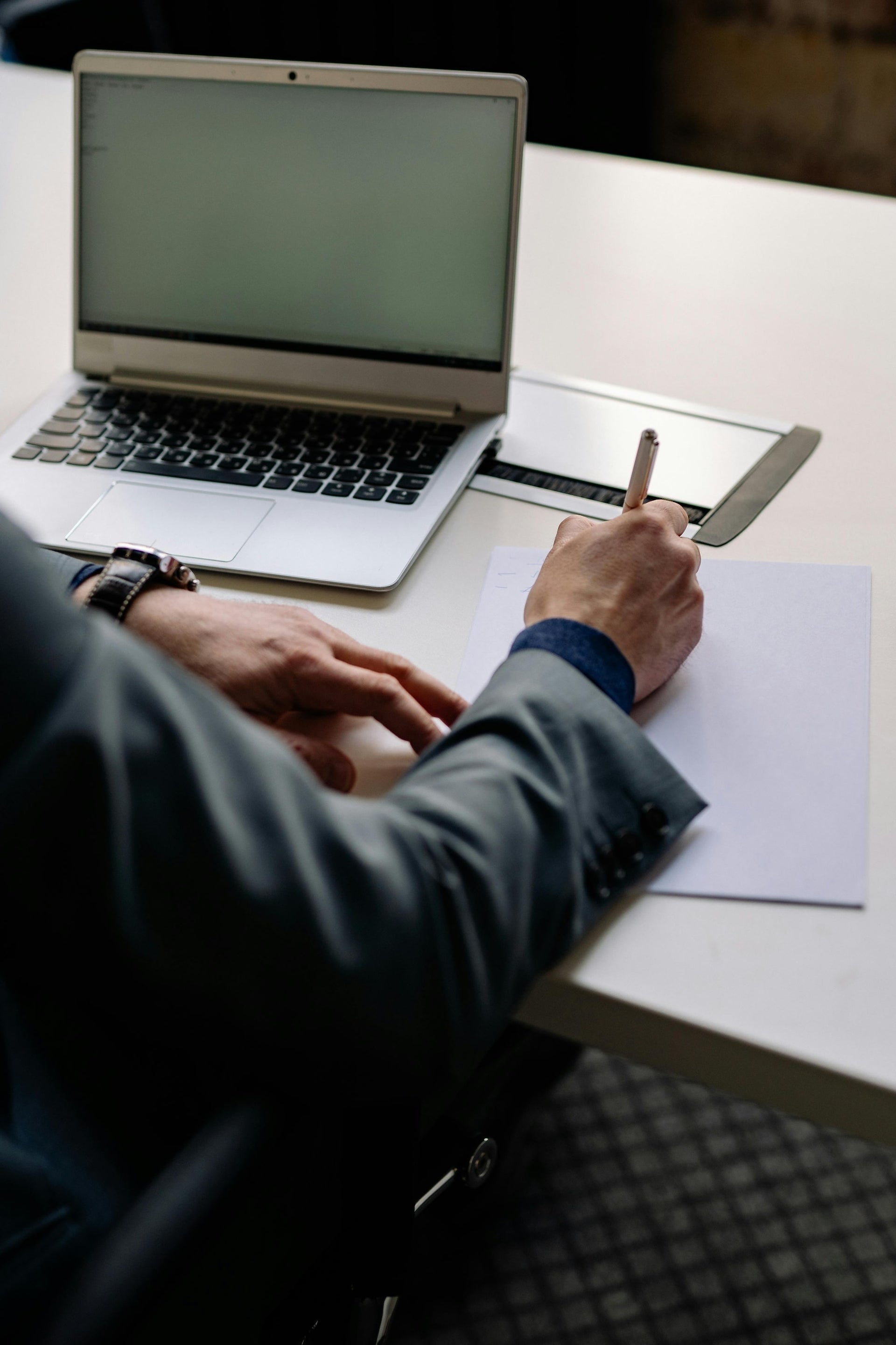 This image shows a person seated at a desk, writing on a sheet of paper with a pen, while a laptop is open in front of them. The individual is dressed in business attire, indicating a professional setting. The scene suggests a combination of digital and handwritten work, highlighting the blend of traditional and modern methods in a business environment.