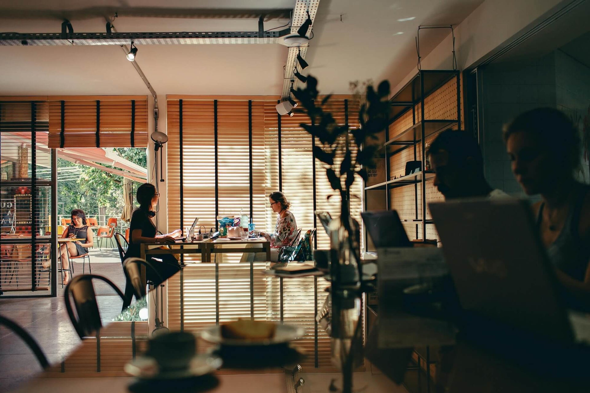 This image captures a cozy cafe scene where people are working on laptops at various tables, with sunlight filtering through wooden blinds. In the foreground, silhouettes of customers are visible, while in the background, a woman is seated outside on a patio, enjoying the relaxed atmosphere. The overall ambiance is warm and inviting, with a mix of natural light and shadow adding to the peaceful environment.