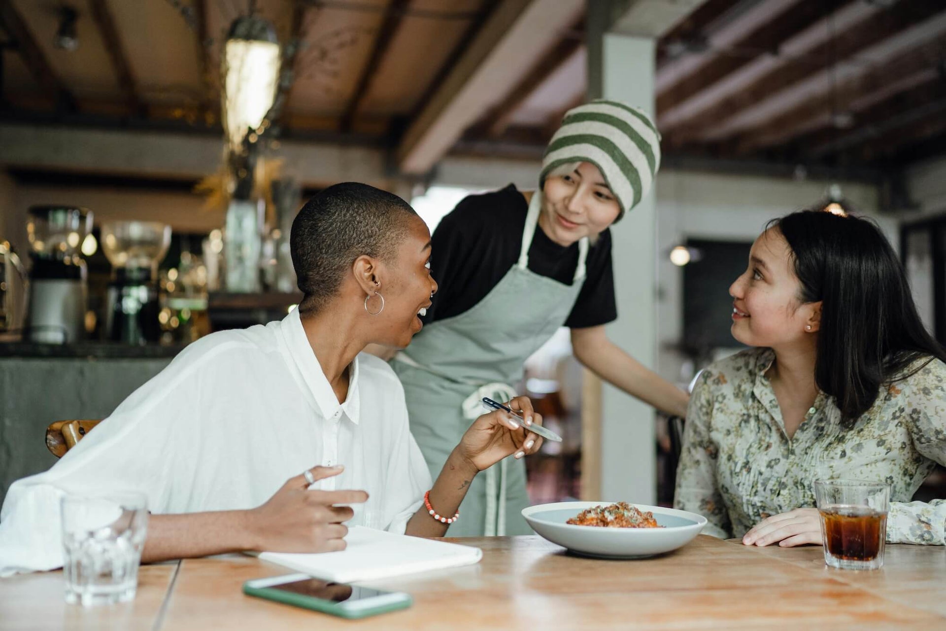 Two women are seated at a table in a restaurant, smiling and engaging in conversation with a friendly waiter wearing an apron and beanie. One of the women has a plate of food in front of her, and both have drinks on the table. The atmosphere is warm and inviting, with a casual and relaxed vibe.