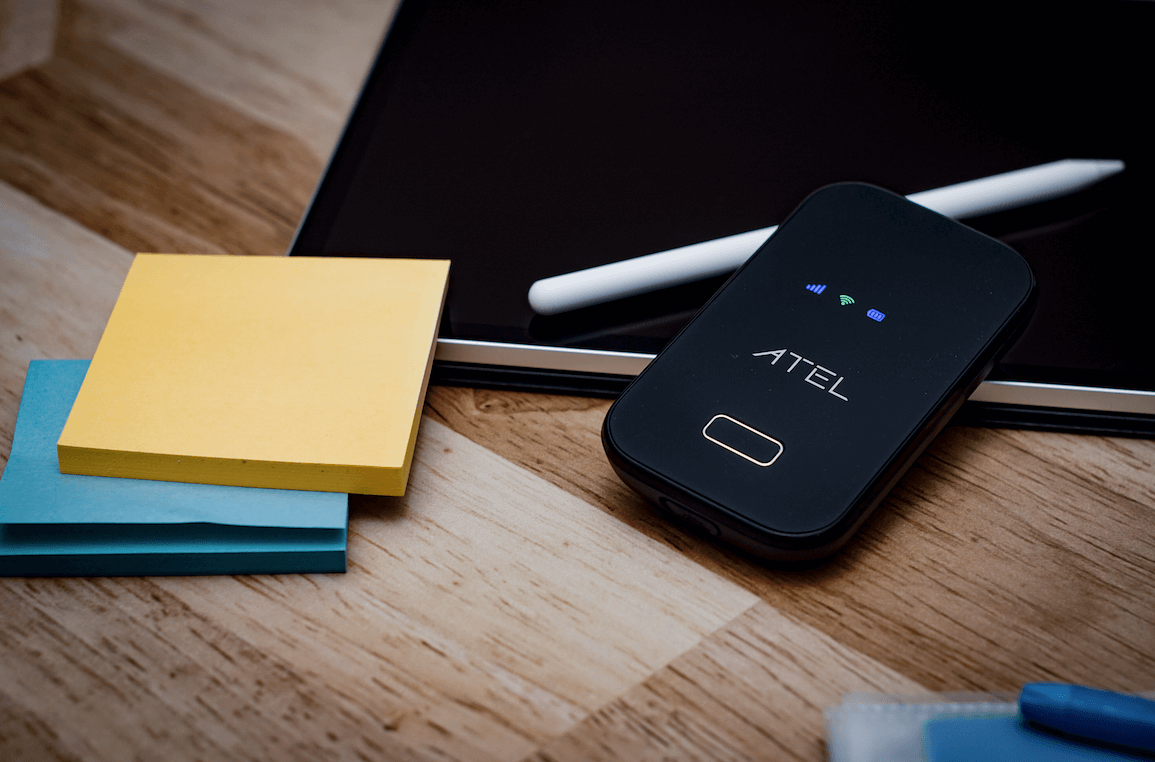 This image features the ATEL W01 mobile hotspot placed on a wooden desk next to a stack of colorful sticky notes, a tablet, and a stylus. The sleek black design of the hotspot is illuminated by small indicator lights, signaling its active connection. The setup suggests a portable, organized workspace, highlighting the convenience and mobility provided by the ATEL W01 device.