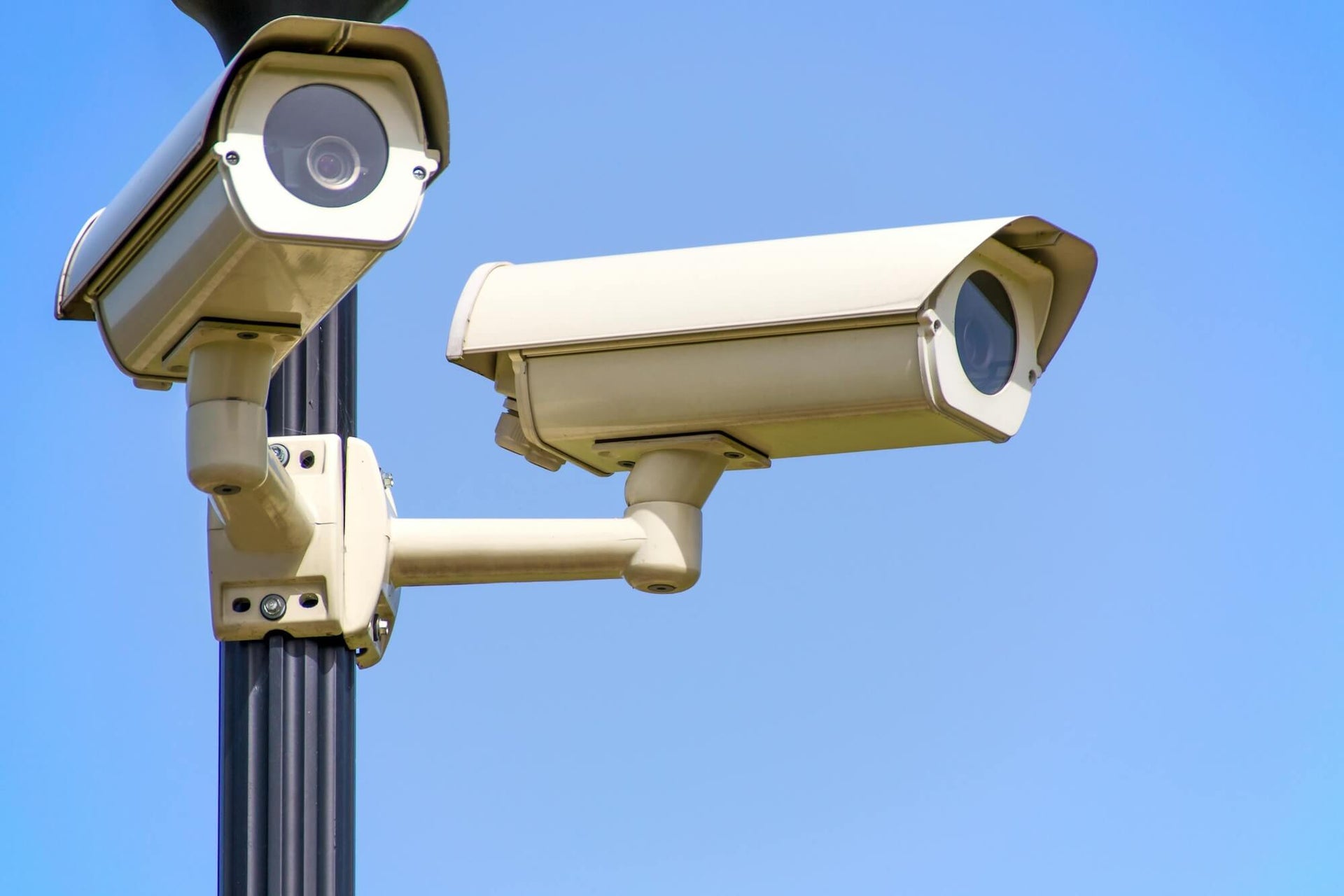 This image shows two security cameras mounted on a pole, angled to monitor different directions. The cameras are beige in color and have a boxy design, typical of outdoor surveillance equipment. They are set against a clear blue sky, emphasizing their presence in an outdoor environment. The image conveys a sense of security and vigilance, as the cameras are positioned to cover a wide area. The overall scene is bright and well-lit, highlighting the cameras as the focal point.