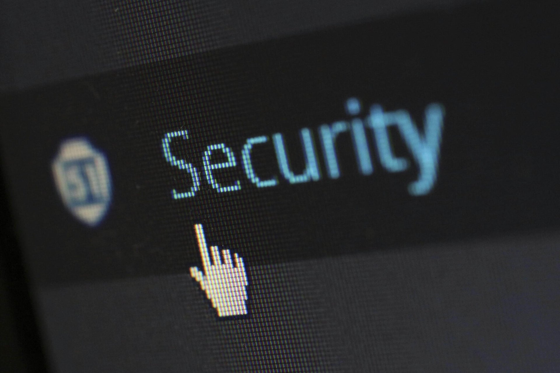 This image shows a close-up of a computer screen displaying the word "Security" with a cursor hovering over it. The screen features a pixelated view, highlighting the text and a shield icon beside it. The image conveys a focus on cybersecurity or digital protection.