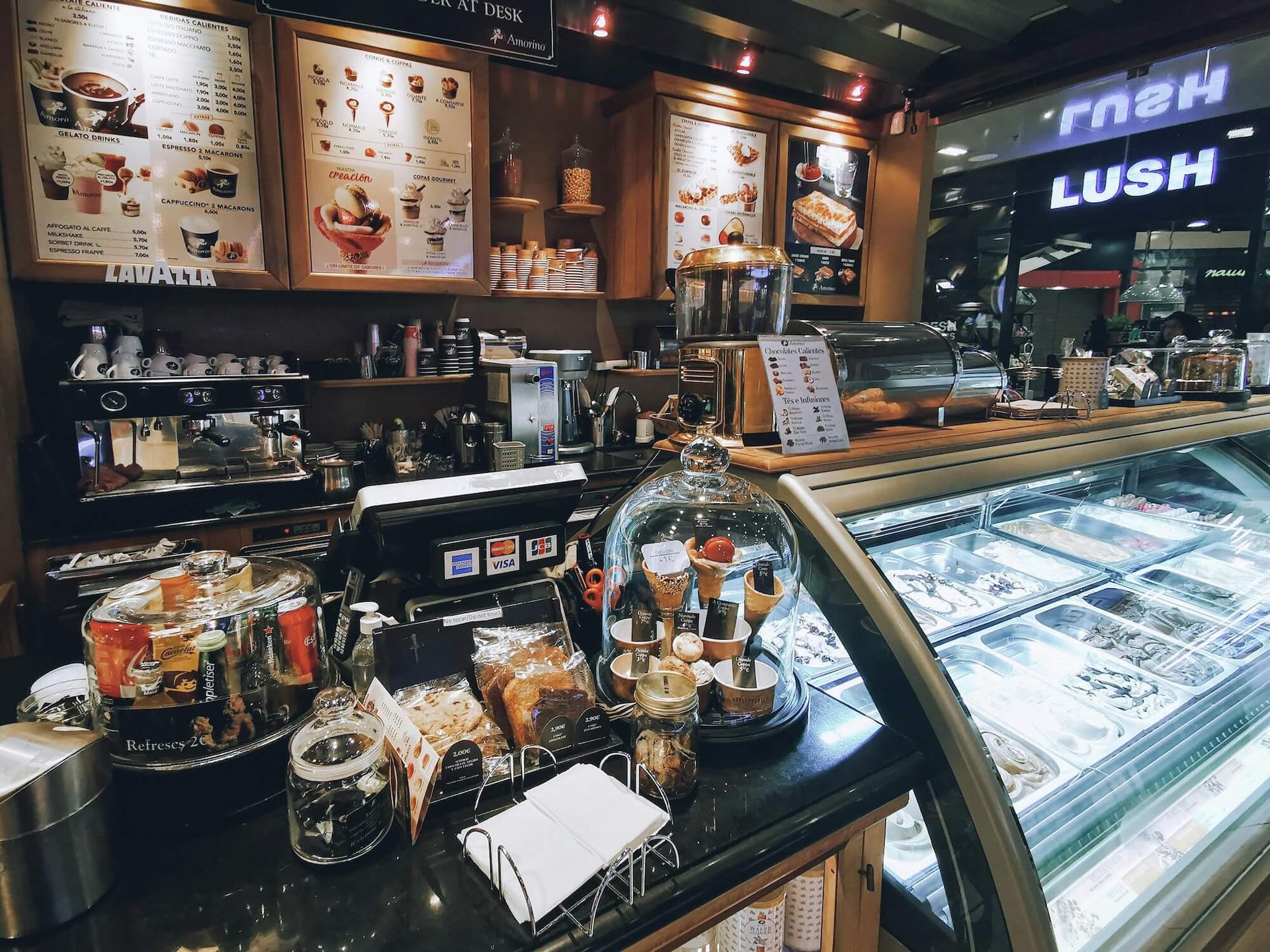 This image shows the interior of a cafe or gelato shop with a well-stocked counter displaying various pastries, snacks, and beverages. Behind the counter, there is an espresso machine, coffee cups, and a menu board featuring different coffee and dessert options. The brightly lit gelato display case showcases multiple flavors, and the overall ambiance is warm and inviting.