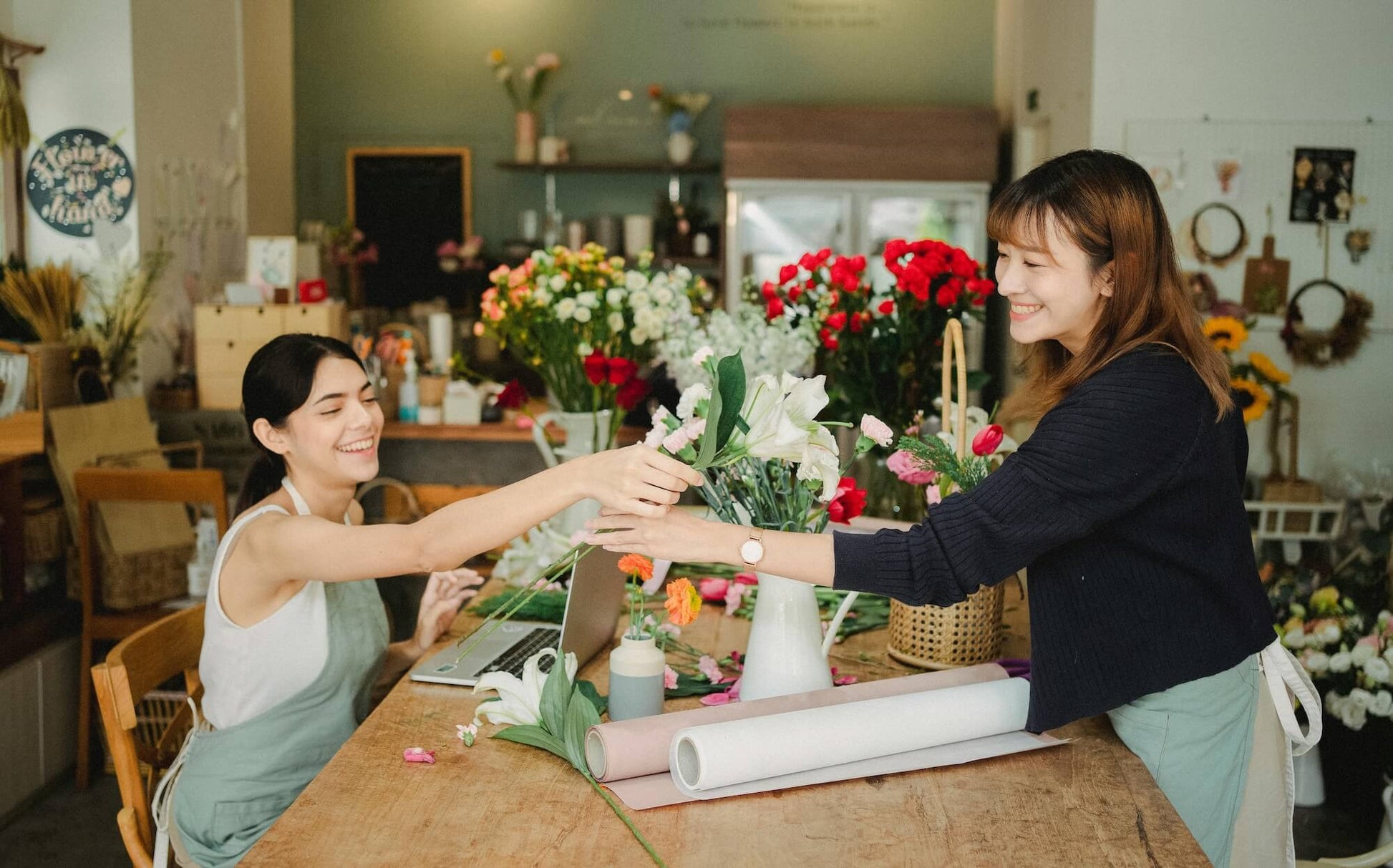 This image shows two women in a flower shop, happily interacting while arranging and handling flowers. One woman, seated at a wooden table, hands a flower to the other woman, who is standing and smiling as she accepts it. The background is filled with a variety of colorful flowers and floral supplies, creating a warm and inviting atmosphere.