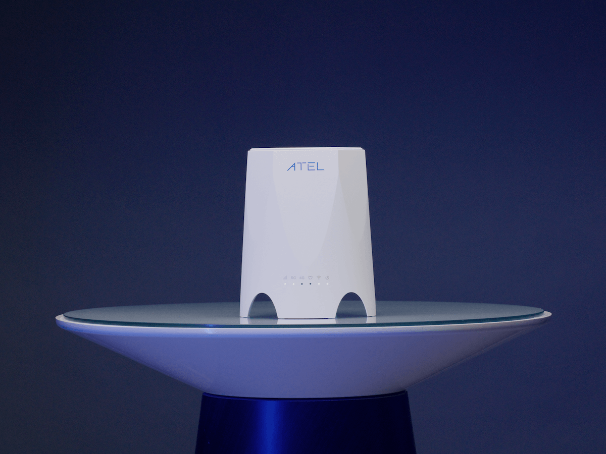 This image showcases the WB550 5G Indoor Router by ATEL. The router is sleek and modern, with a white exterior and minimalist design. It is placed centrally on a glass-topped surface with a blue base, against a solid dark blue background, emphasizing the product's clean lines and contemporary appearance. The front of the router features subtle indicator lights and the ATEL logo near the top, highlighting its brand. The overall image has a professional and high-tech feel, making the product the focal point.