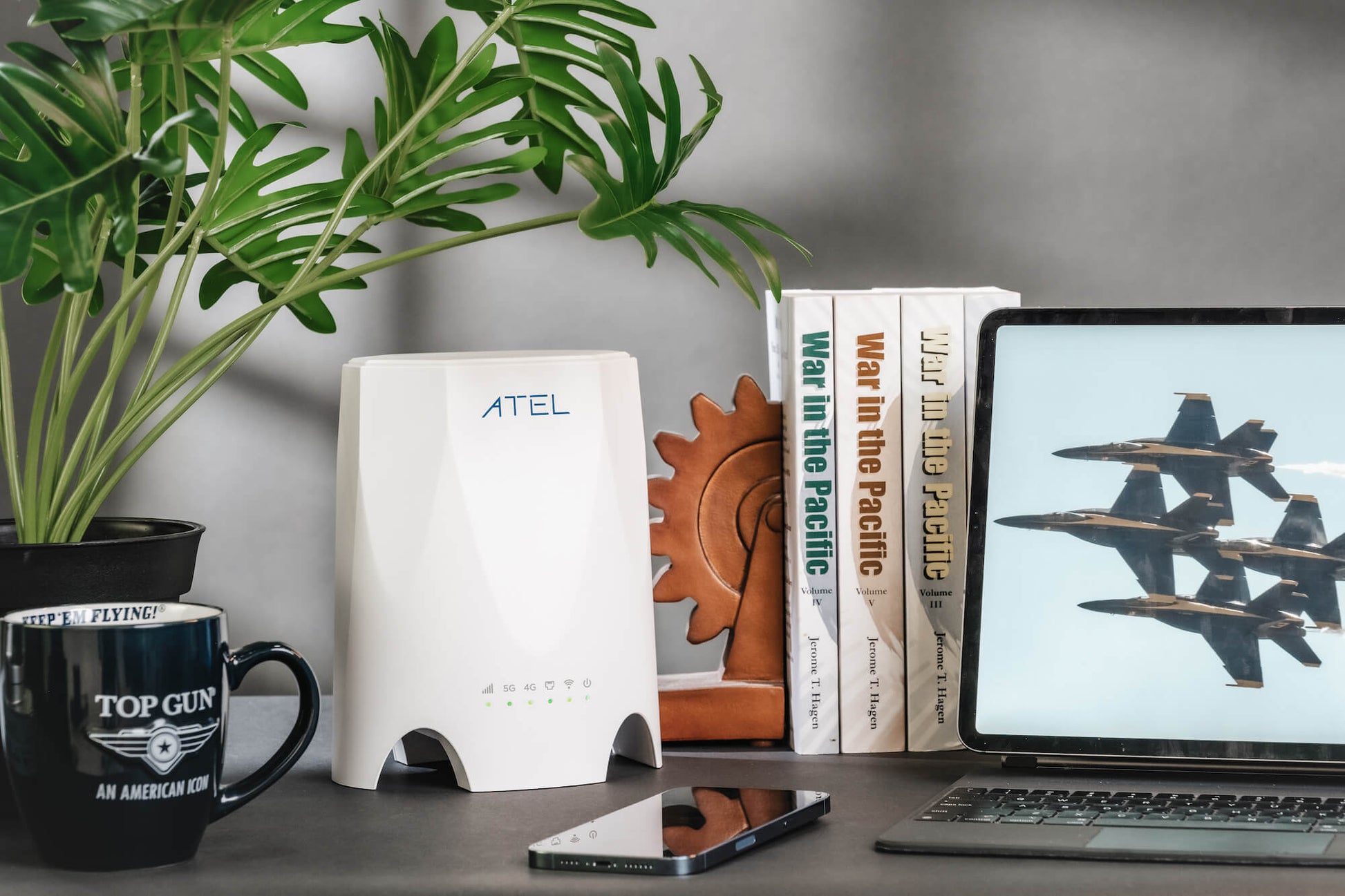 This image features a workspace with a sleek white WB550 5G Fixed Wireless Access Router prominently displayed on a desk. The router is positioned next to a laptop showing a picture of jet fighters in flight, a smartphone, and a stack of books titled "War in the Pacific." The scene is completed with a potted plant and a "Top Gun" coffee mug, suggesting a high-performance and technology-driven environment.